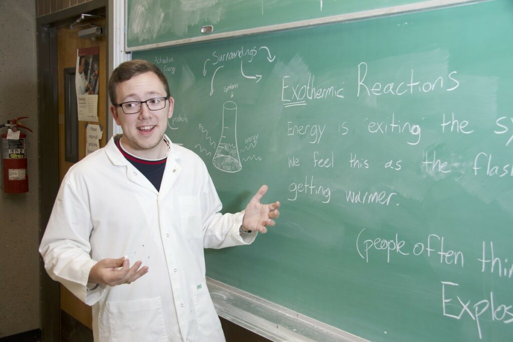 A man in a lab coat speaks while writing on a chalk board