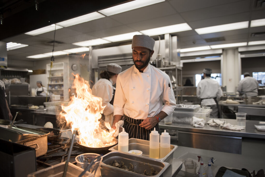 A student in a chef uniform holds a frying pan, practicing to flambe.