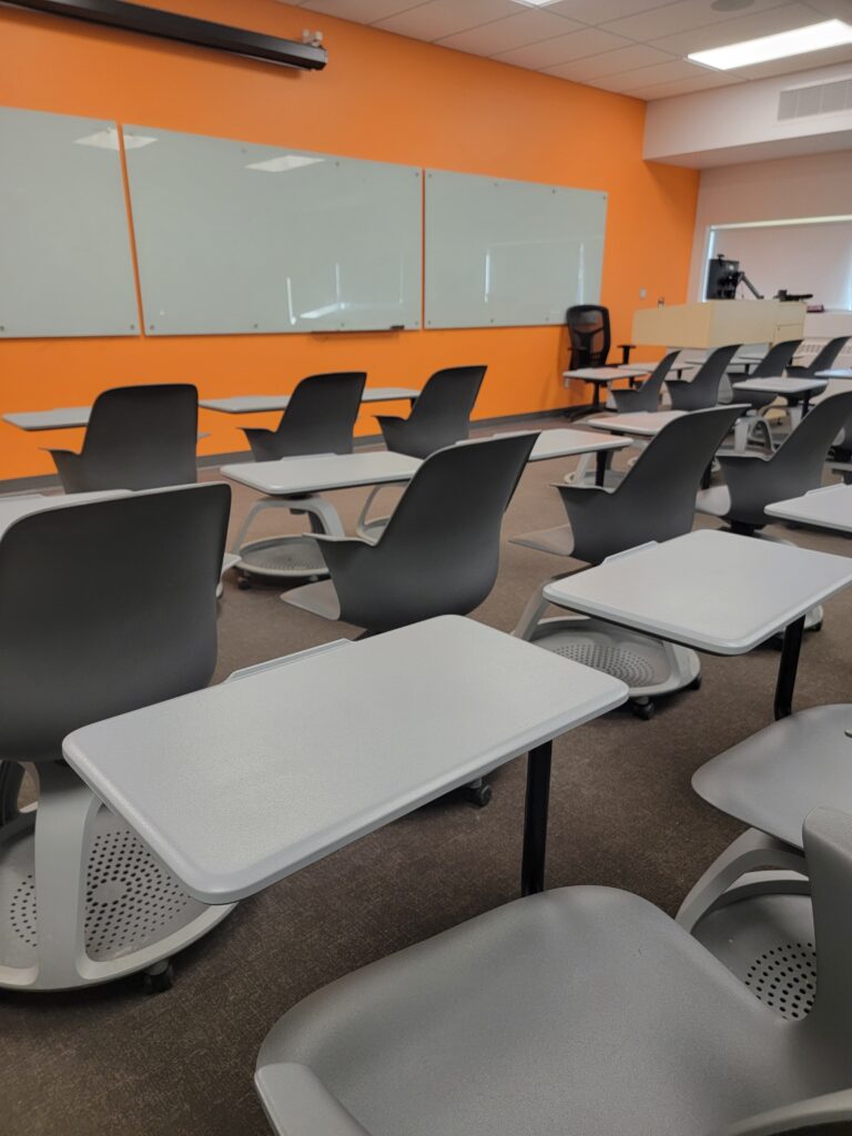 Photo of free-standing chairs on wheels with desks attached in front of some whiteboards.