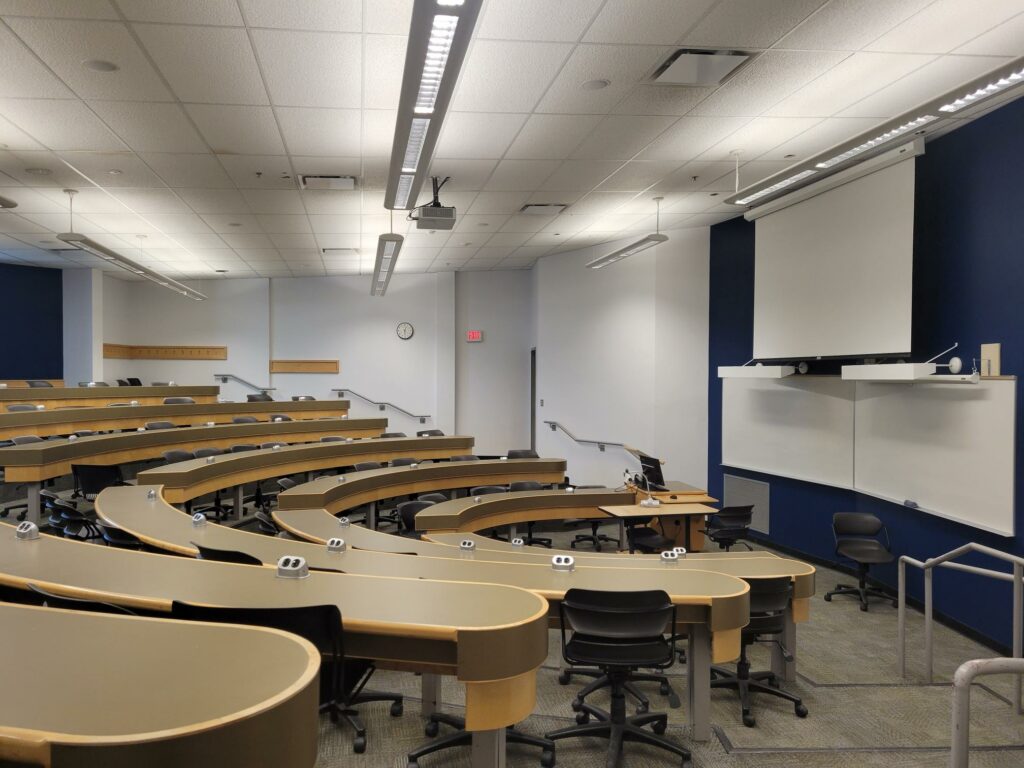 Photo of a lecture room with long tables and desk chairs.