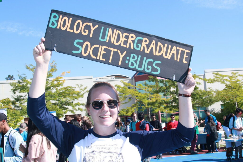 A smiling student holds up a sign that says "Biology Undergraduate Society: BUGS"