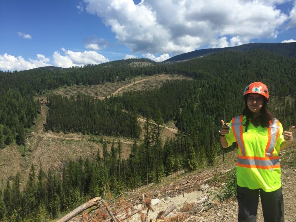A student stands on a hillside in a hard hat and safety vest giving a thumbs-up.
