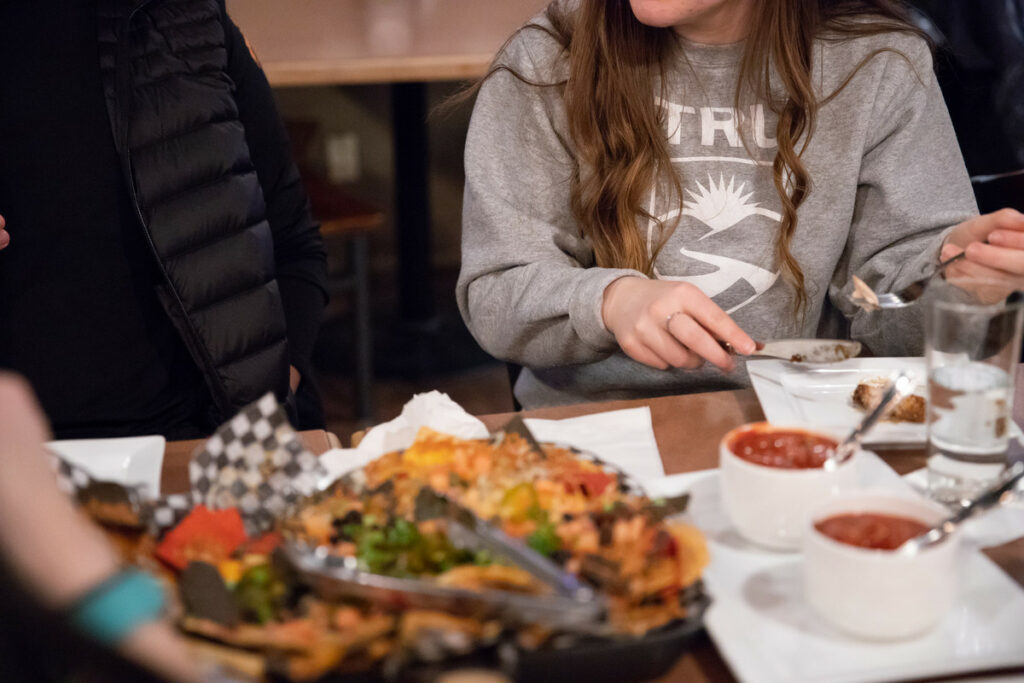 A woman in a TRU sweater eats in a pub next to someone in a black puffer jacket. There is a plate of nachos and salsa in front of them.