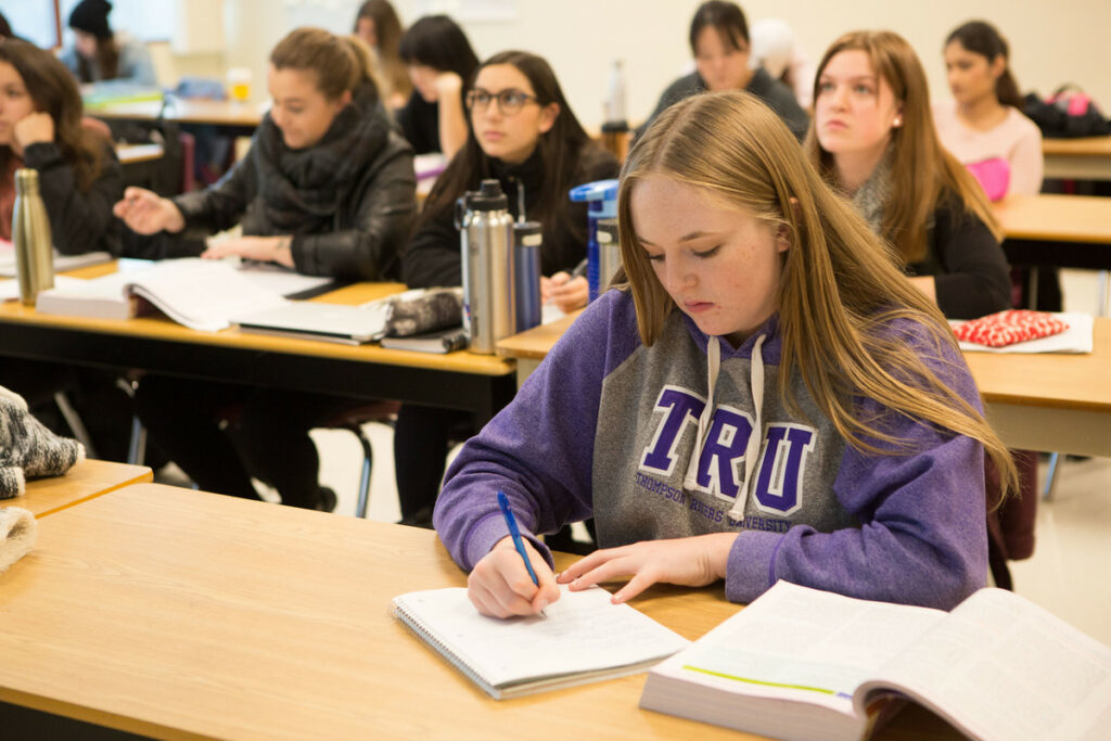A student in a TRU hoodie writes in a notebook text to a textbook during a lecture.