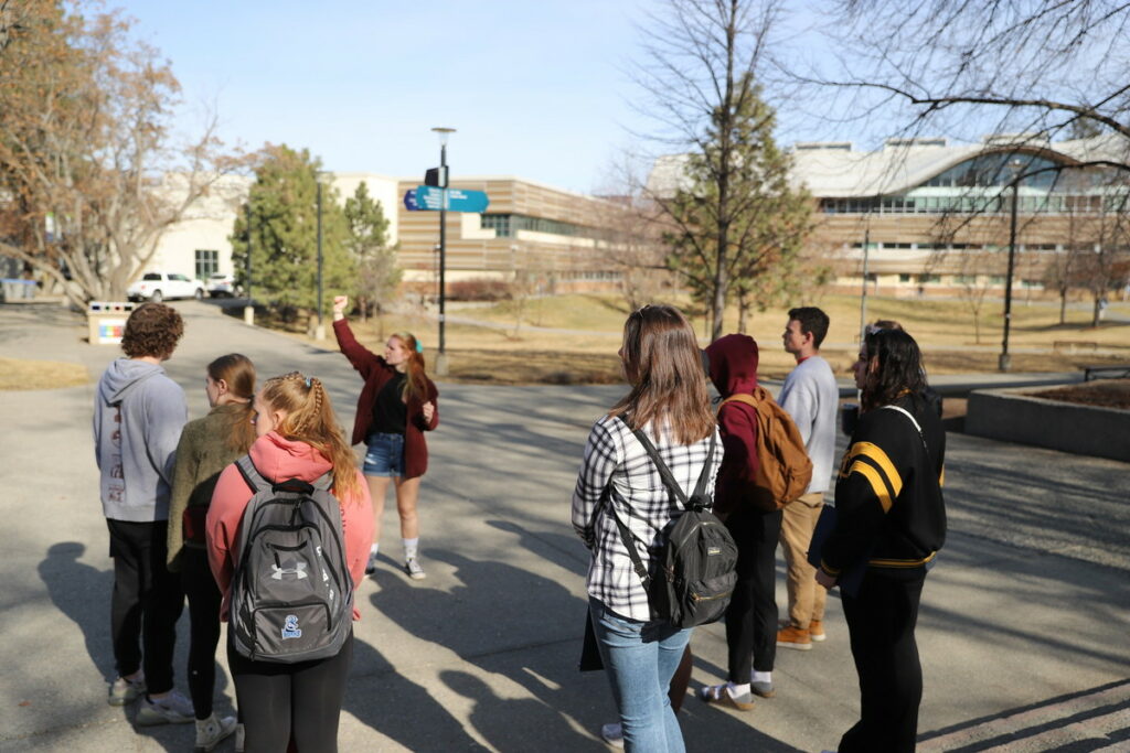 A group of 7 people listen while a tour guide gestures to a building during a campus tour.