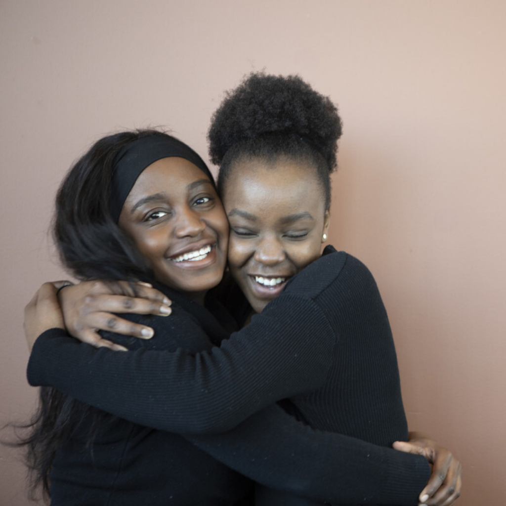 Two girls in black sweaters hug while smiling and facing the camera.